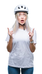 Young blonde woman wearing cyclist security helmet over isolated background amazed and surprised looking up and pointing with fingers and raised arms.