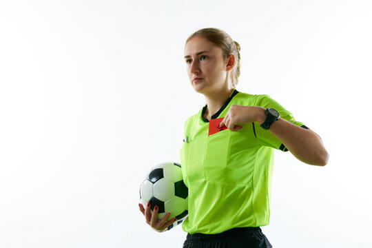 Young woman, female soccer referee holding ball, stopping game and showing red card against white studio background. Concept of sport, competition, match, profession, football game, control