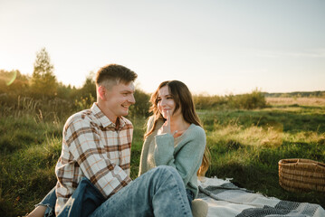Couple sitting on blanket, hugging in grass in field at sunset. Happy young woman and man, walking spending time together in nature. Concept of family holiday outdoors. Female embrace male on picnic.