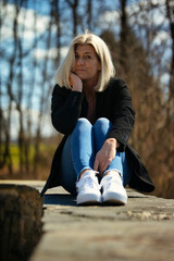 Woman in her mid-fifties with blonde long hair in a portrait, dressed in a black jacket, white T-shirt, blue jeans and white sneakers, outside in autumnal nature.