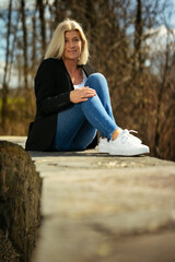 Woman in her mid-fifties with blonde long hair in a portrait, dressed in a black jacket, white T-shirt, blue jeans and white sneakers, outside in autumnal nature.