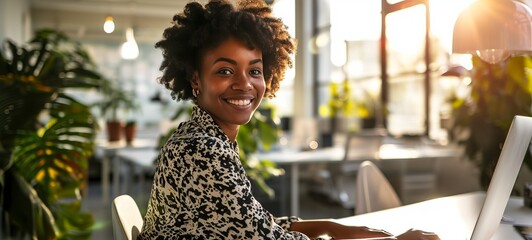 Close-up portrait of a young African American woman with curly hair sitting in a bright office or coworking space with a laptop. Beautiful smiling girl works or studies with enthusiasm.
