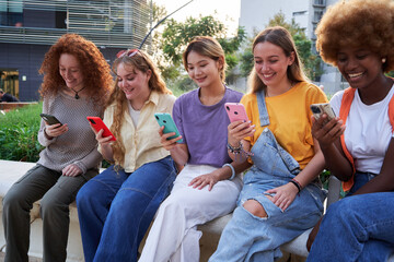 Multiracial group of young women friends enjoying and smiling using their phones sitting in the...