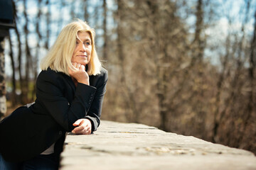 Woman in her mid-fifties with blonde long hair in a portrait, dressed in a black jacket, white T-shirt, blue jeans and white sneakers, outside in autumnal nature.