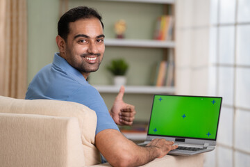 Happy indian man using green screen laptop by showing thumbs up while looking at camera at home - concept of satisfaction, approval and advertisement