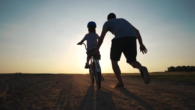 family play in the park. father teaching daughter to ride a bike. happy family kid dream concept. daughter learn to ride a bike silhouette. father supporting child riding bike summer in sunset park