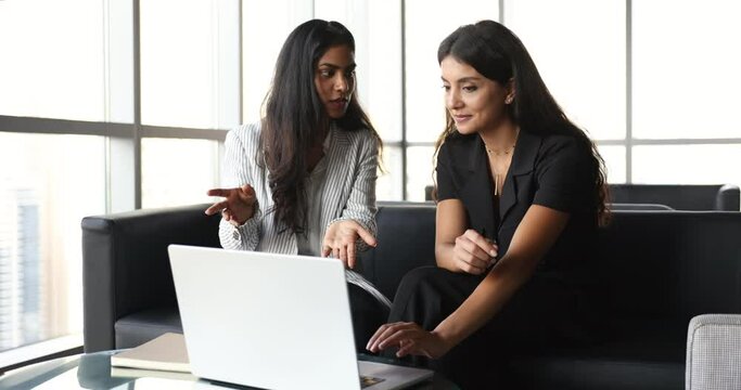Two attractive multiethnic women colleagues using laptop met in modern skyscraper office, look at computer screen, watch online presentation, make improvements, prepare joint task, engaged in teamwork