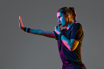 Athletic pose, young woman referee blowing whistle with serious expression against grey studio background in neon light. Concept of sport, competition, match, profession, football game, control - obrazy, fototapety, plakaty
