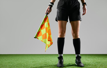Cropped image of female legs, referee on field standing with flag to signal game rules against grey studio background. Concept of sport, competition, match, profession, football game, control - obrazy, fototapety, plakaty