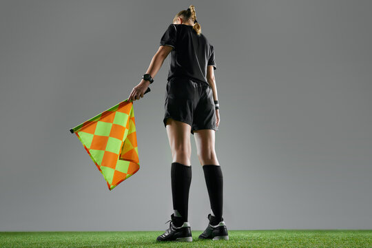 Bottom back view. Young woman, female referee in uniform standing with flag, controlling game rules against grey background. Concept of sport, competition, match, profession, football game, control