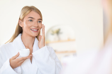 Attractive blonde young lady applies moisturizer in modern bathroom