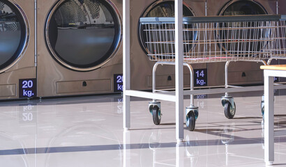 Stainless steel laundry basket trolley near table set on tile floor with row of vending washing machines and clothes dryer inside of modern laundromat shop - Powered by Adobe
