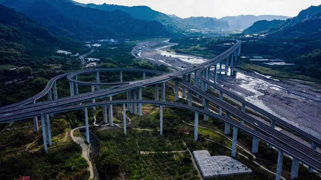 hyperlapse, time-lapse photography, drone aerial view, highway interchange intersection, daytime. the sun beam passes through the clouds, flicker, river and mountain landscape behind it. taiwan