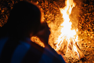 Bright bonfire at night in nature, backshot of a girl sitting near it and drinking warm tea. Concept of spending time outdoors in nature, enjoying the burning of bonfire