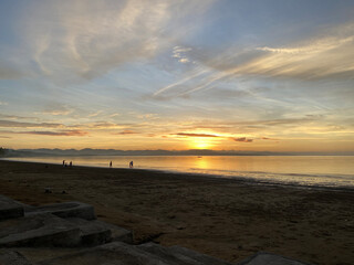 On the beach, the golden early sunrise casts clouds on the sea horizon. Unrecognizable figures enjoy the view. Silhouettes