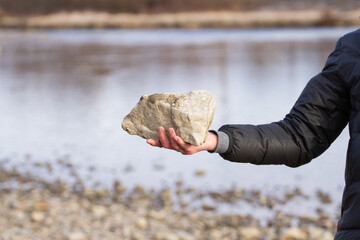 A man holds a large stone on the bank of a river in spring.