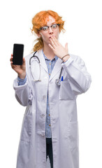 Young redhead doctor woman showing smartphone cover mouth with hand shocked with shame for mistake, expression of fear, scared in silence, secret concept