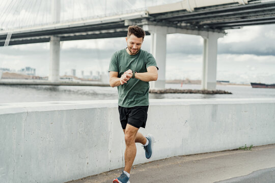 Focused man checking his sports watch while jogging by the city waterfront. - Powered by Adobe