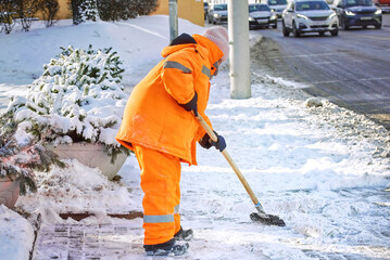 Utility worker in orange uniform breaking ice with ice breaker tool, remove ice with steel razor scraper on sidewalk at bus stop. Clean and remove ice and snow with icebreaker tool. Snow removal work - obrazy, fototapety, plakaty