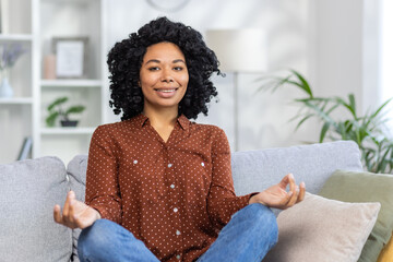 Portrait of a smiling young African American woman sitting on the couch at home in the yoga lotus...