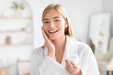 Blonde woman in bathrobe applies face cream in light bathroom