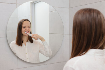 Toothbrush technology trends. Maintaining a bright smile. Daily dental care routine. Home appliances for oral health. Caucasian young adult woman brushing teeth in bathroom in front of mirror