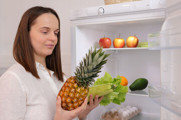 Delighted cute Caucasian woman standing near open refrigerator full of fruits and vegetables takin fresh pineapple and green lettuce leaves thinking what to cook