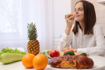 Satisfied delighted Caucasian brown haired woman wearing white shirt sitting at table in kitchen with fruit and vegetables smelling fresh apple cooking healthy meal