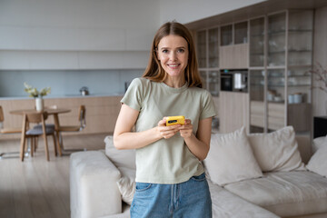 Portrait smiling woman looking at camera holding smartphone at home. Happy charming girl standing in living room with mobile phone. Dreaming relaxed young female resting at home. Good mood, daytime.