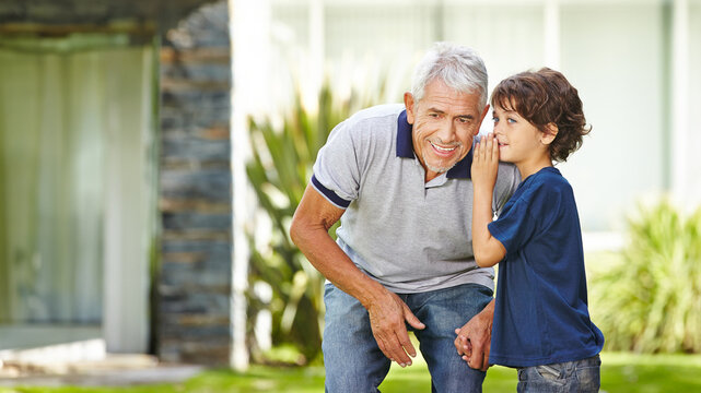 Grandson whispering secret in ear of grandfather at home in garden