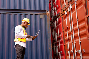 black african man wears hardhat and vest holding laptop computer checking container cargo,young...