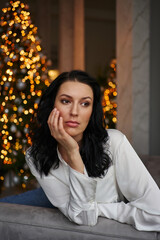 Fototapeta premium Gracefully seated in a grey chair, the young woman in a white shirt captivates against the backdrop of sparkling lights from a Christmas tree.