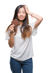 Young asian woman eating chocolate bar over isolated background stressed with hand on head, shocked...