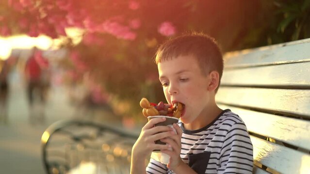 Boy sits on a bench and eats ice cream wrapped in a waffle with chocolate and strawberries