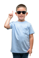 Dark haired little child wearing sunglasses surprised with an idea or question pointing finger with happy face, number one