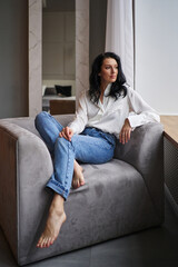 Nestled by the window in a chic setting, a brunette beauty donned in a white shirt and jeans graces a grey chair, epitomizing contemporary allure.