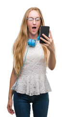 Blonde teenager woman holding smartphone and wearing headphones scared in shock with a surprise face, afraid and excited with fear expression