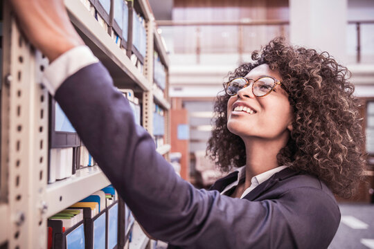 Young African corporate woman looking through shelves with binders and books in large start-up office space. Cape Town, South Africa