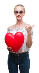 Blonde teenager woman holding red heart pointing and showing with thumb up to the side with happy face smiling