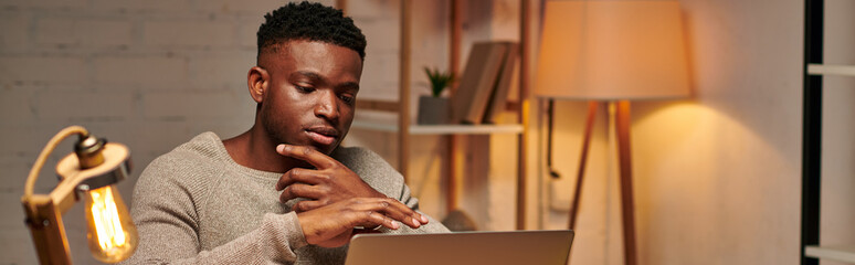 creative african american freelancer thinking near laptop while working from home at night, banner