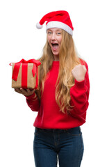 Blonde teenager woman wearing santa claus hat screaming proud and celebrating victory and success very excited, cheering emotion