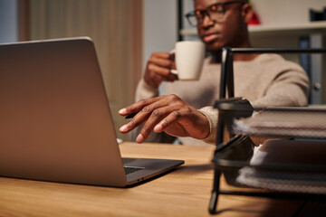 young african american freelancer with coffee cup working at laptop in home office at night