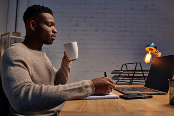 african american guy with coffee cup writing in notebook near laptop working in home office at night