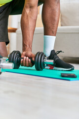 Fototapeta na wymiar partial view of fit african american man in sportswear training with dumbbell in living room at home