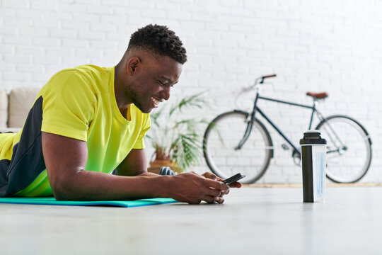 joyful sporty african american man with smartphone lying down on fitness mat near sports bottle - Powered by Adobe