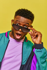 impressed african american man with open mouth holding sunglasses and looking at camera on yellow - obrazy, fototapety, plakaty