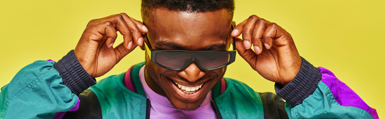 trendy african guy in bright jacket adjusting sunglasses and smiling on yellow backdrop, banner