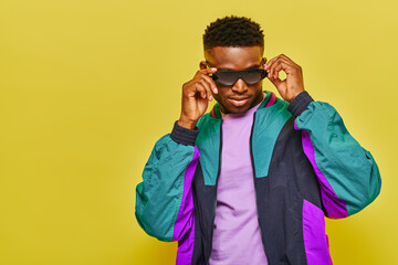 expressive african american man in trendy windbreaker jacket adjusting stylish sunglasses on yellow