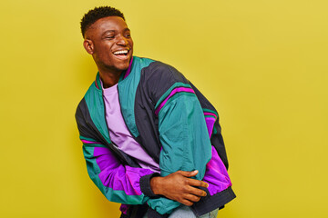 excited african american man in bright windbreaker jacket laughing and looking away on yellow