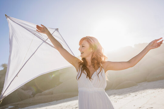 Beautiful woman with red hair in white dress dancing and having fun on the beach with kite. Cape Town, South Africa